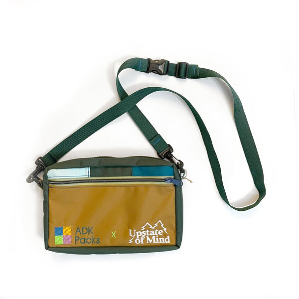 An Adirondack Packs and Upstate of Mind bag collab. Featuring a crossbody fanny pack with various earthy toned swatch fabrics, and embroidered "ADK Packs x Upstate of Mind" text.