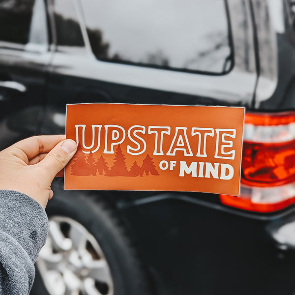 Upstate of Mind Bumper Sticker | Someone holding up an orange background with darker orange trees and tan text that reads "Upstate of Mind", in front of a black SUV car.