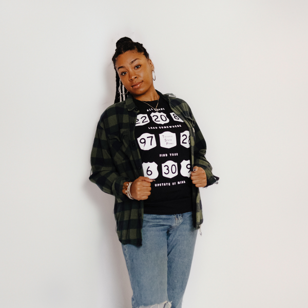 A model wearing our All Roads Tee in Black Heather with a green plaid flannel and blue jeans.