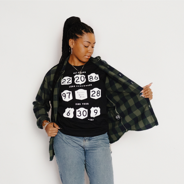 A model wearing our All Roads Tee in Black Heather with a green plaid flannel and blue jeans.