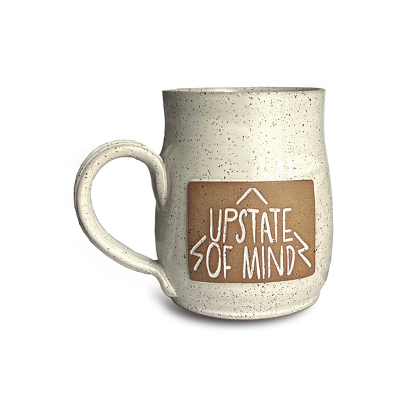 A handthrown, handmade ceramic mug by Folk and Wayfarer Pottery. Speckled white and tan with a clay square that reads "Upstate of Mind" on the front of the mug. Very nice and unique!