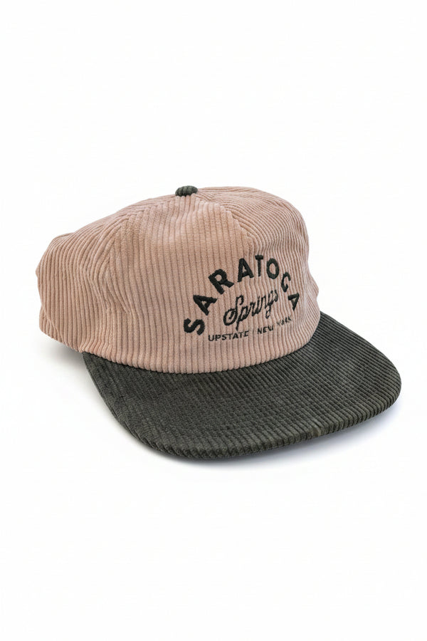 A corduroy baseball cap with a beige crown and a dark green brim, featuring 'SARATOGA Springs' embroidered on the front. The cap is resting on a dark tabletop with other items in the background.