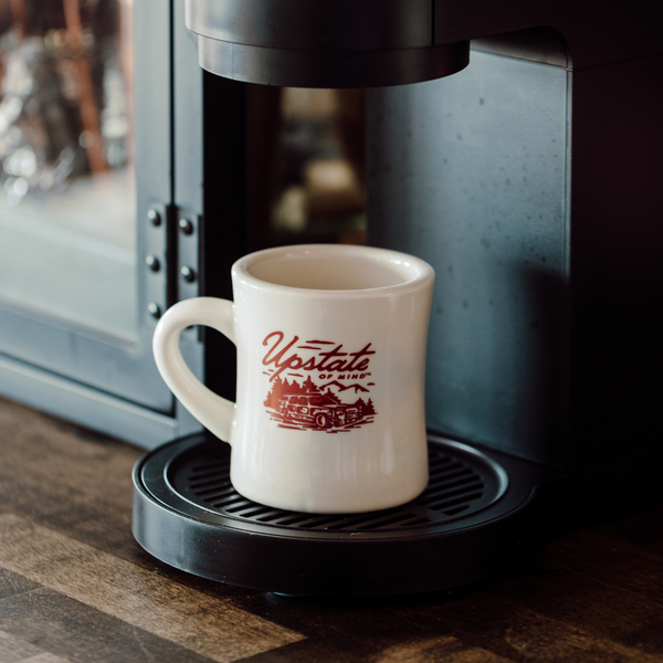 A lifestyle photo of The Woody Wagon Ceramic Mug. White background, the mug is a light creme, tan color, and the graphic is burgundy with a design of a vintage station wagon in the wood. Text on the mug reads "Upstate of Mind", and is under a Keurig coffee machine.