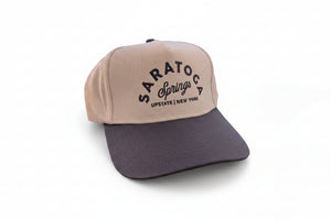 A two-tone baseball cap with a cream-colored front panel and a dark gray brim, featuring 'SARATOS SPRINGS UPSTATE NEW YORK' printed in black on the front. The cap is resting on a dark green table with a wire basket of folded yellow shirts in the background.