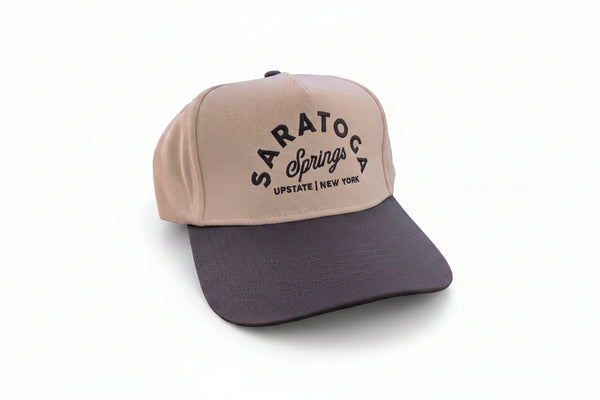 A two-tone baseball cap with a cream-colored front panel and a dark gray brim, featuring 'SARATOS SPRINGS UPSTATE NEW YORK' printed in black on the front. The cap is resting on a dark green table with a wire basket of folded yellow shirts in the background.