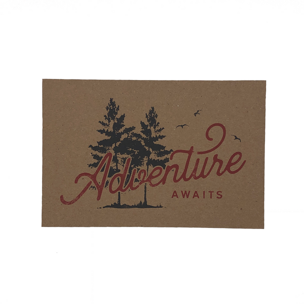 Photo of "ADVENTURE AWAITS" postcard with a silhouette of ink trees and bird. Printed on brown cardboard paper.