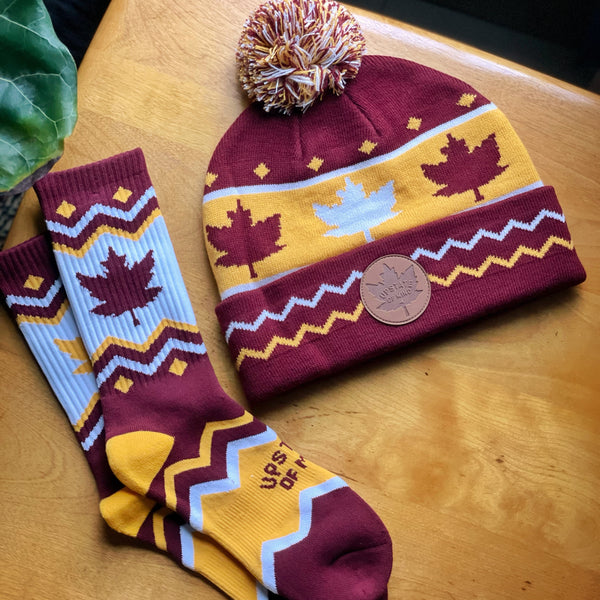 Upstate of Mind Maple Leaf Socks. Colors are burgundy and yellow with chevron stripes, blocks of yellow/burgundy, a yellow maple leaf print, and text that reads "Upstate of Mind". Photo is of the socks and matching maple beanie.