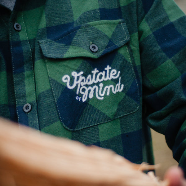UOM Chain Stitch Woven Button-Up Flannel - Forest Plaid. Close-up of the text on front left chest pocket that is in script and reads "Upstate of Mind".