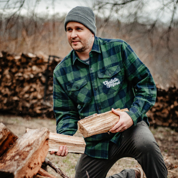 Model wearing UOM Chain Stitch Woven Button-Up Flannel - Forest Plaid while collecting firewood. Text on the front left chest pocket is in script and reads "Upstate of Mind".