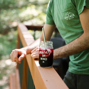 A lifestyle photo of our Upstate of Mind Mason Jar Pint Glass. Model is enjoying an iced cold berry drink in the woodsy summer.